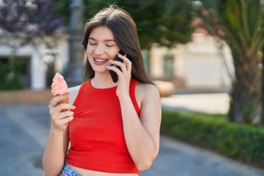 Beyaz bir kadın akıllı telefondan konuşuyor. Parkta dondurma yiyor.