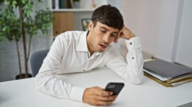 Tükenmiş genç İspanyol bir adam, çekici bir profesyonel, iş yerinde sıkılmış ve yorgun, akıllı telefonunun ekranına bakıyor, kendini ofiste sıkışmış hissediyor.