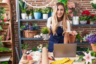 Young blonde woman florist using laptop holding plant at florist