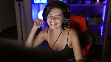 Kendine güvenen genç İspanyol bir kadın, güzel bir flama, oyuna dalmış, karanlık oyun odasında yayılıyor, teknoloji parlıyor, kulaklık takıyor, neşeli bir gülümseme geceyi aydınlatıyor.