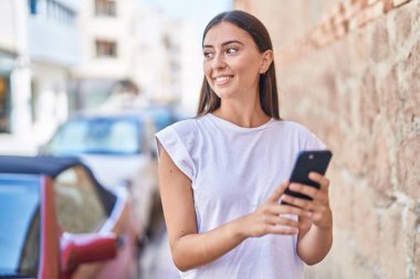 Genç, güzel, İspanyol bir kadın sokakta akıllı telefon kullanarak gülümsüyor.