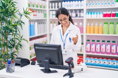 Young beautiful hispanic woman pharmacist using computer holding pills bottle at pharmacy