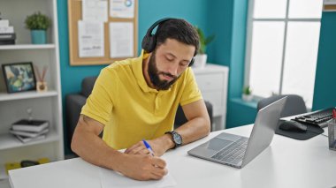 Genç İspanyol iş adamı. Laptop ve kulaklık kullanıyor. Ofiste not alıyor.