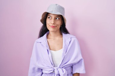 Pembe arka planda duran genç İspanyol kadın. Şapka takmış, gülümsüyor, yan tarafa bakıyor ve düşünüyor.. 