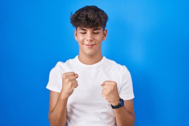 Latin kökenli genç, mutlu ve heyecanlı bir şekilde kollarını kaldırarak, gülümseyerek ve başarı için bağırarak zafer jesti yapıyor. kutlama konsepti. 