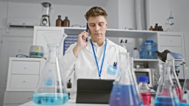 Yakışıklı beyaz erkek bilim adamı ciddi ciddi akıllı telefondan konuşuyor, modern, yoğun laboratuarda dokunmatik alanda geziniyor, önemli bir teknolojik buluşa odaklanmış durumda..