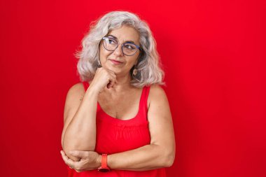 Orta yaşlı bir kadın, kırmızı arka planda duran gri saçlı, elleri çenesinde soru soran, düşünceli bir yüz ifadesi olan. Gülümseyen ve düşünceli bir yüz. şüphe kavramı. 