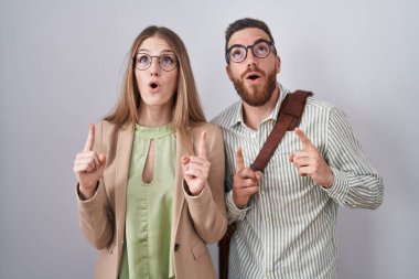 Beyaz arka planda duran genç çift şaşkınlık ve şaşkınlık içinde yukarı bakıyor ve parmaklarıyla işaret ediyor ve kollarını kaldırıyordu.. 