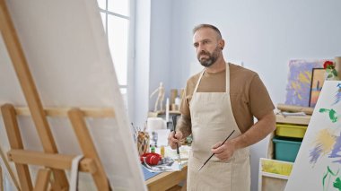 Yakışıklı genç sanatçı, rahatlamış sanat atölyesinde yoğun bir şekilde resim çiziyor. Boya fırçaları, tuvaller ve konsantre yetişkin öğrencilerle dolu bir yaratıcılık sınıfı..