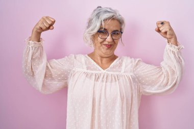 Middle age woman with grey hair standing over pink background showing arms muscles smiling proud. fitness concept. 