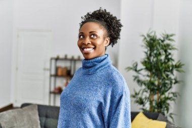 Afro-Amerikalı bir kadın gülümseyerek evde duruyor.