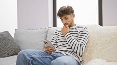 Ciddi genç Arap adam bir sonraki fikrinden şüphe ediyor, akıllı telefonuyla mesajlaşırken derinden dalmış, oturma odasındaki kanepede tek başına oturuyor, basit bir an, online teknoloji denizinde..