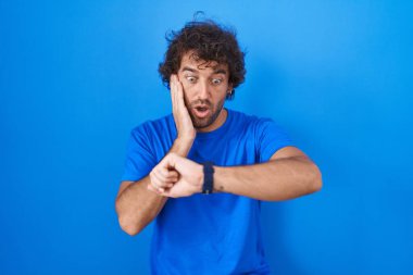 Hispanic young man standing over blue background looking at the watch time worried, afraid of getting late 