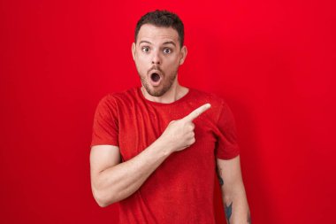 Young hispanic man standing over red background surprised pointing with finger to the side, open mouth amazed expression. 