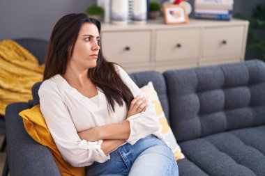 Young beautiful hispanic woman sitting on sofa with serious expression at home
