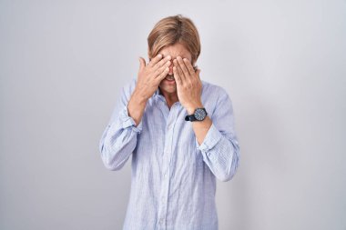 Beyaz arka planda duran beyaz bıyıklı beyaz bir adam yorgunluk ve baş ağrısı için gözlerini ovuşturuyor, uykulu ve yorgun bir ifade takınıyor. görme sorunu 