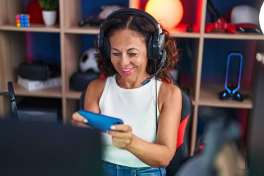 Middle age woman streamer playing video game using smartphone at gaming room