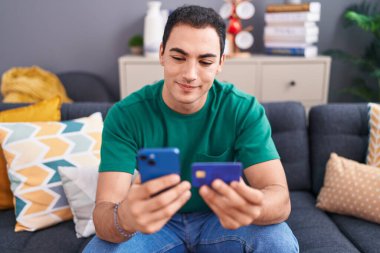 Genç İspanyol adam akıllı telefon ve kredi kartı kullanıyor. Evdeki kanepede oturuyor.