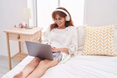 Middle age woman using laptop and headphones sitting on bed at bedroom