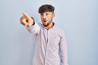 Mavi arka planda duran sakallı bir Arap, parmağını ileri uzatmış, ağzı açık, şaşırmış, ön tarafta bir şey var. 