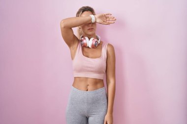 Spor giyim ve kulaklık takan genç sarışın kadın gözlerini kollarıyla kapatıyor, ciddi ve üzgün görünüyor. Görme, saklama ve reddetme konsepti 