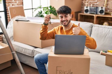 Yeni ev anahtarlarını sakallı İspanyol bir adam elinde dizüstü bilgisayarlı, mutlu ve pozitif gülümseyen bir şekilde arıyor. 