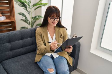 Young beautiful hispanic woman psychologist writing on document at psychology clinic