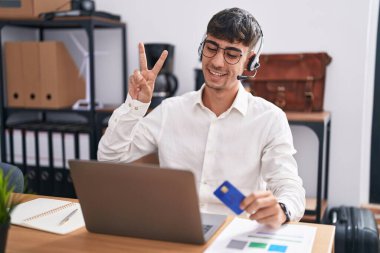 Bilgisayarda çalışan İspanyol genç adam elinde kredi kartı ile kameraya gülümseyen ve parmaklarıyla zafer işareti yapan. İki numara.. 