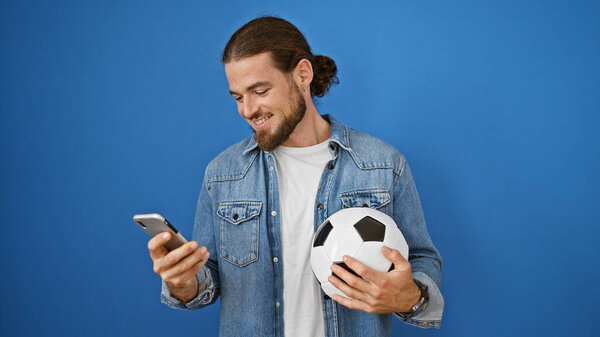 Young hispanic man smiling holding soccer ball and smartphone over isolated blue background