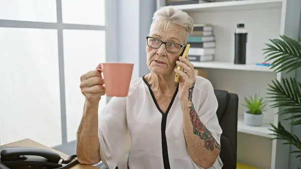 エレガントな灰色の髪の女性の上司は深刻な電話会話に魅了され 彼女のオフィスワークステーションでリラックスしてコーヒーを飲む — ストック写真