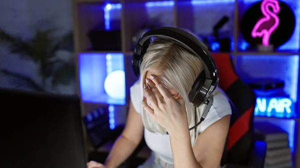 Overworked and stressed young blonde female streamer battling stress while gaming late night in a gloomy home office