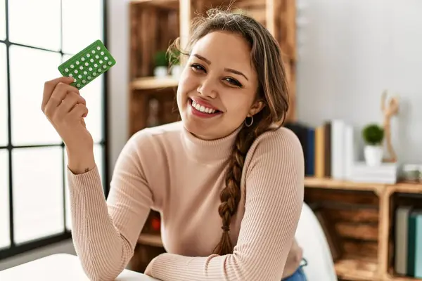 Young beautiful hispanic woman holding birth control pills sitting on table at home