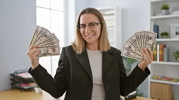 Beautiful young blonde business woman standing, confidently holding and counting dollars at her workspace in the office. a successful professional boss emanates happiness and wealth.