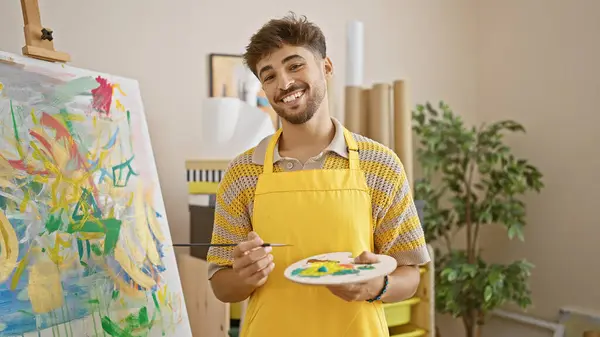 Smiling young arab man showcasing his paintbrush talent at art studio, confidently embracing his painting hobby and education