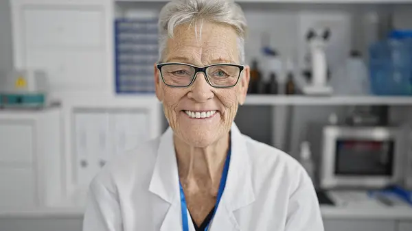 自信と笑顔の灰色の髪の女性科学者は 彼女の仕事を楽しんで 顕微鏡とテストチューブの中でラボに幸せに座って — ストック写真
