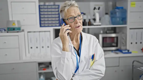 賑やかな実験室活動の中で彼女のスマートフォンで真剣に話している灰色の髪のシニア科学者女性 — ストック写真