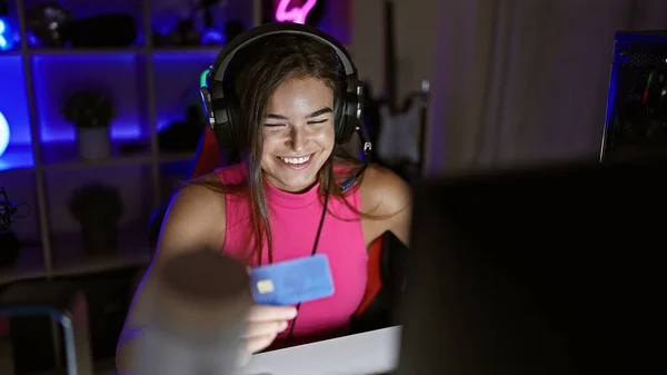 Beautiful young hispanic streamer, smiling confidence radiating, buys gaming gear with credit card amidst nightly streaming session in her gaming room