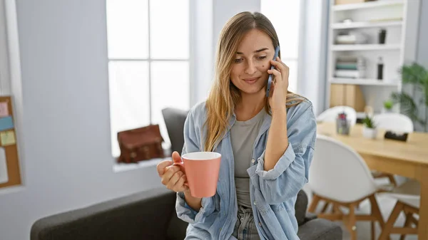 Vibrant, confident blonde boss woman sparking success with smartphone talk & enjoying espresso in elegant office interior