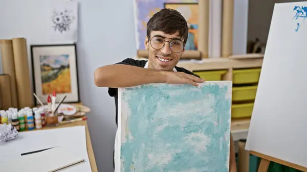 Smiling, confident young hispanic man draws passionately at art studio, a portrait of a handsome, smiling artist with paintbrushes in hand, basking in the warmth of creativity.
