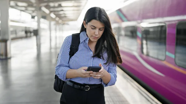 Young beautiful hispanic woman walking down the railway using smartphone at train station