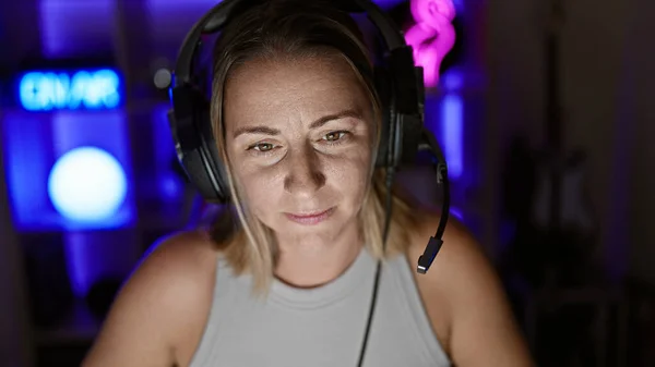 Attractive young blonde woman streamer, locked in her gaming room, focused and serious, using futuristic technology whilst live streaming video game play on her computer at night
