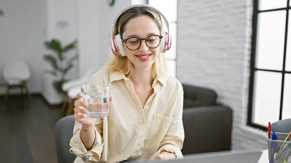 Young blonde woman business worker using laptop and headphones drinking water at the office