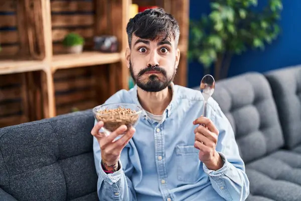 Young hispanic man with beard eating healthy whole grain cereals puffing cheeks with funny face. mouth inflated with air, catching air.
