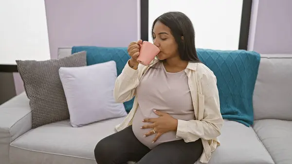 Home comfort, young pregnant woman enjoying a relaxing morning at home, drinking coffee and touching her belly on a comfortable sofa