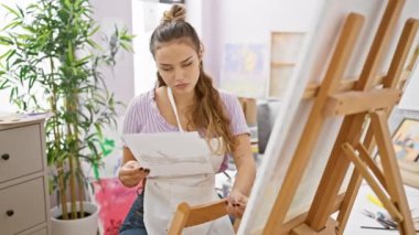 Meraklı genç İspanyol kadın sanatçı, sanatına iyice dalmış, bir stüdyoda oturuyor, önlük giymiş, elinde boya fırçası, resmine dalgın dalgın dalgın bakıyor..