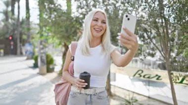 Neşeli genç sarışın kadın güvenli bir şekilde sıcak kahvesinin tadını çıkarırken parkta parlak bir şekilde gülümsüyor, şehrin güneş ışığını emiyor, telefonuyla çevrimiçi bir video görüşmesine bağlanıyor..