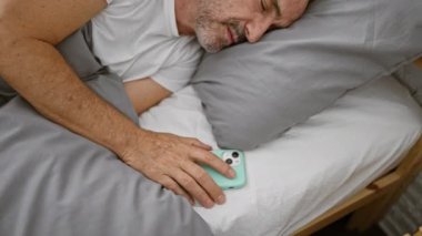 Yorgun, orta yaşlı, gri saçlı, rahat yatak odasında dinlenen, pijamalarıyla yatağa uzanmış, akıllı telefonunu tutarken derin bir uykuya dalmış bir adam..