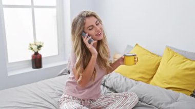 Mutlu genç bir kadın, yatakta akıllı telefonuyla rahatça konuşurken kahvesini yudumluyor. Sabahları rahat yatak odasında dinleniyor.