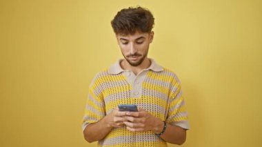 Neşeli genç Arap adam kendine güveni tam, akıllı telefonuyla zaferini kutluyor, izole edilmiş sarı bir arka plana geniş bir gülümseme ile bakıyor.