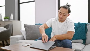 Yakışıklı genç Çinli adam, rahat ama konsantre olmuş, evdeki kanepede otururken bilgisayarını açıyor. Asyalı bir yetişkinin oturma odasında online çalışmak için teknolojiyi kullanırken çekilmiş bir fotoğrafı..